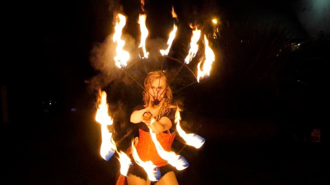 A performer holds two semi-circular fire pois in each hand. Each of the four tines of the pois hold a flaming torch. The pois are held in such a way that the woman is framed by a flaming circle.